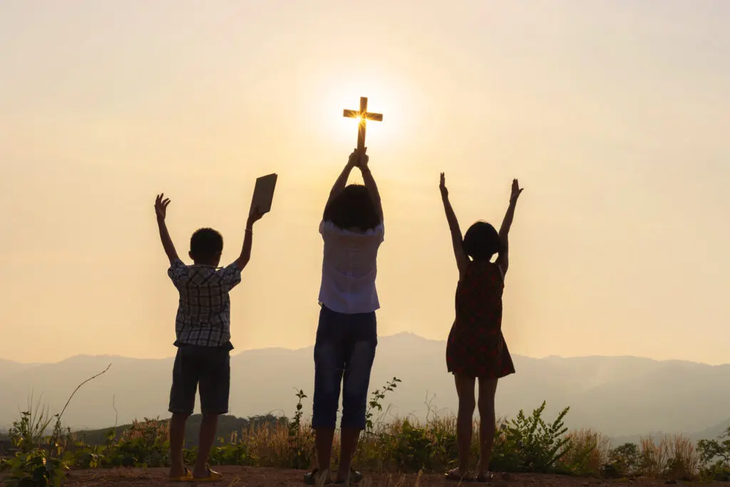 Silhouette of children praying to the god while holding a crucifix symbol with bright sunbeam on the sky